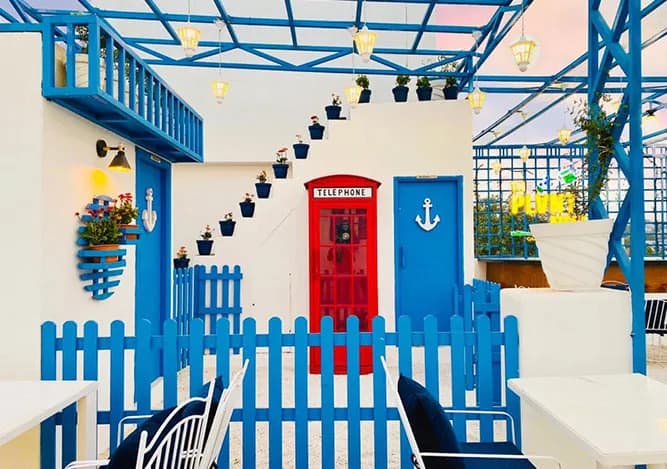 White rooftop courtyard with a blue picket fence, blue doors, a red telephone-box door and potted plants